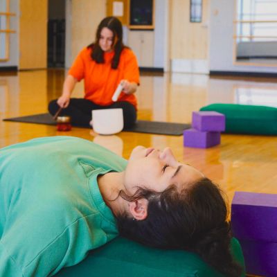 A person lays flat on the floor while a person in the background plays soothing sounds on a singing bowl